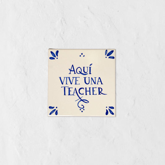 Azulejo decorativo de cerámica blanca con bordes y letras pintadas a mano en azul cobalto. En el centro se lee la frase en español 'AQUÍ VIVE UNA TEACHER'. El diseño incluye detalles ornamentales en las esquinas y pequeños puntos decorativos, sobre un fondo de pared blanca con textura rugosa.