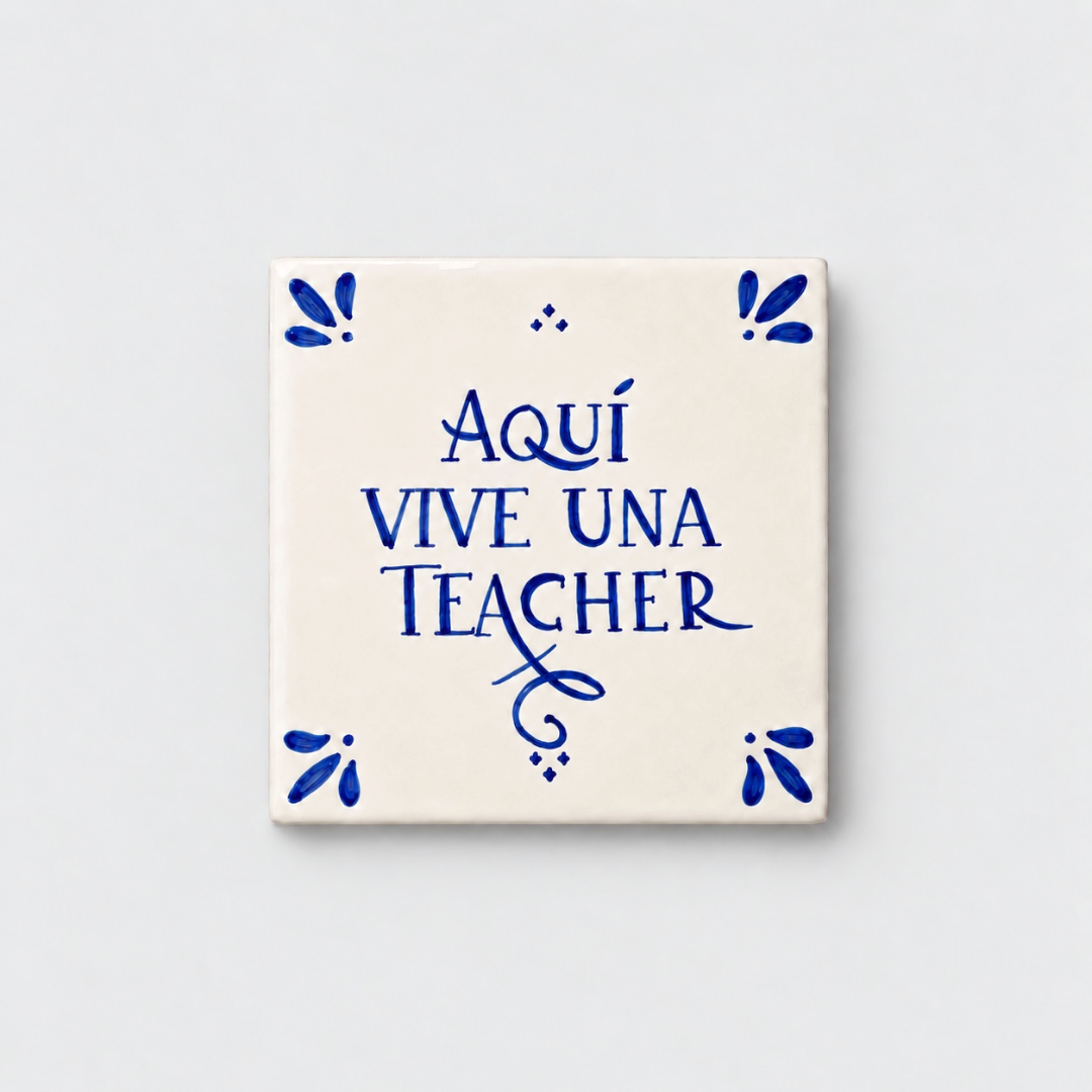 Azulejo decorativo de cerámica blanca con bordes y letras pintadas a mano en azul cobalto. En el centro se lee la frase en español 'AQUÍ VIVE UNA TEACHER'. El diseño incluye detalles ornamentales en las esquinas y pequeños puntos decorativos, sobre un fondo de pared blanca con textura rugosa.