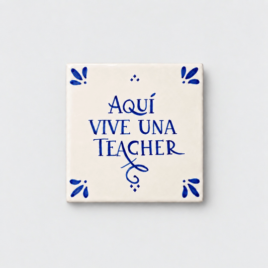 Azulejo decorativo de cerámica blanca con bordes y letras pintadas a mano en azul cobalto. En el centro se lee la frase en español 'AQUÍ VIVE UNA TEACHER'. El diseño incluye detalles ornamentales en las esquinas y pequeños puntos decorativos, sobre un fondo de pared blanca con textura rugosa.