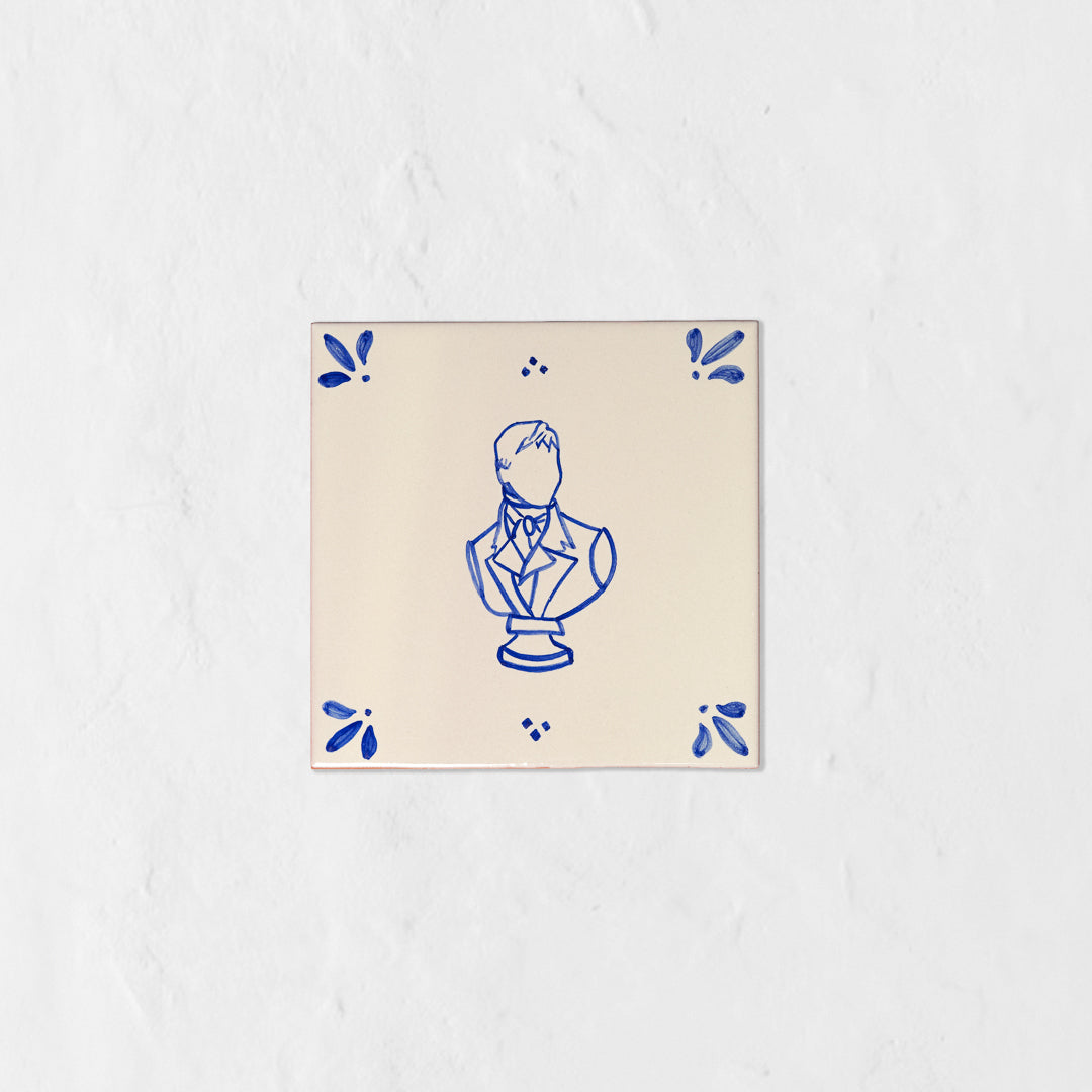 Azulejo decorativo de cerámica blanca con bordes y letras pintadas a mano en azul cobalto. En el centro tiene una ilustración del busto de Mr Darcy. El diseño incluye detalles ornamentales en las esquinas y pequeños puntos decorativos, sobre un fondo de pared blanca con textura rugosa.
