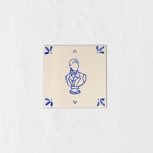 Azulejo decorativo de cerámica blanca con bordes y letras pintadas a mano en azul cobalto. En el centro tiene una ilustración del busto de Mr Darcy. El diseño incluye detalles ornamentales en las esquinas y pequeños puntos decorativos, sobre un fondo de pared blanca con textura rugosa.