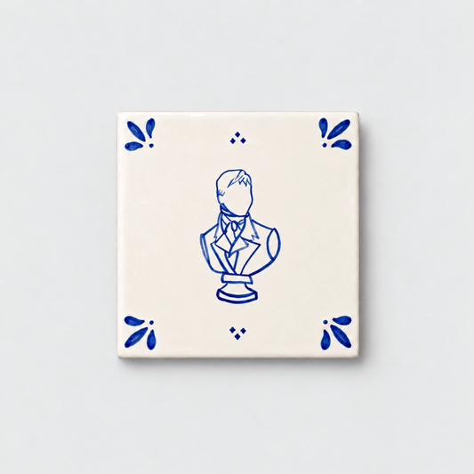 Azulejo decorativo de cerámica blanca con bordes y letras pintadas a mano en azul cobalto. En el centro tiene una ilustración del busto de Mr Darcy. El diseño incluye detalles ornamentales en las esquinas y pequeños puntos decorativos, sobre un fondo de pared blanca con textura rugosa.