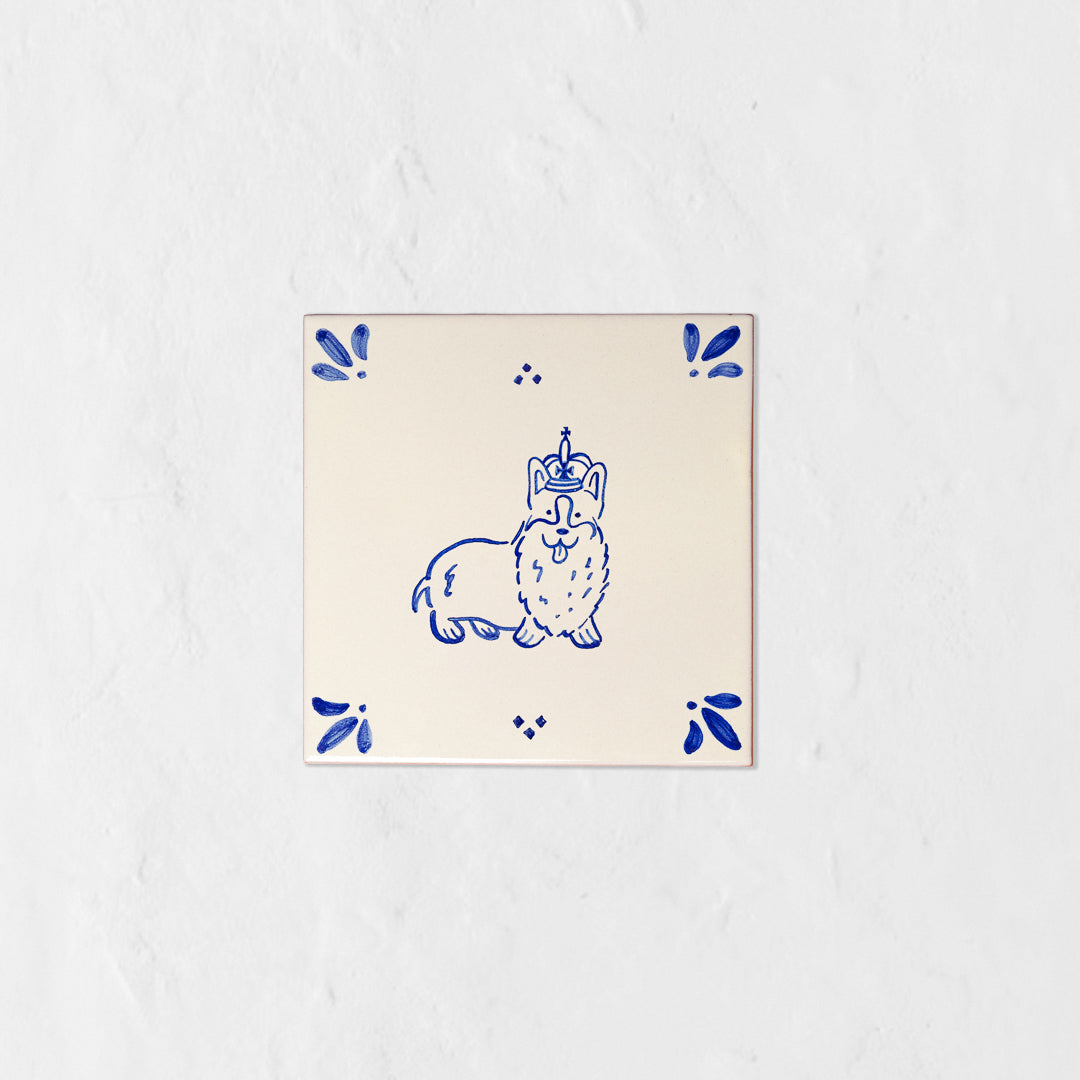 Azulejo decorativo de cerámica blanca con bordes y letras pintadas a mano en azul cobalto. En el centro lleva la ilustración de un corgi con corona. El diseño incluye detalles ornamentales en las esquinas y pequeños puntos decorativos, sobre un fondo de pared blanca con textura rugosa.
