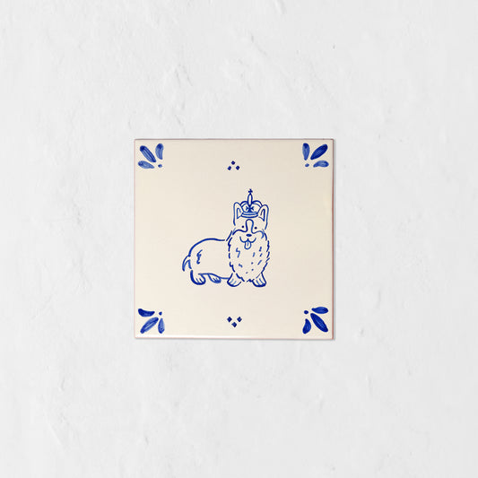Azulejo decorativo de cerámica blanca con bordes y letras pintadas a mano en azul cobalto. En el centro lleva la ilustración de un corgi con corona. El diseño incluye detalles ornamentales en las esquinas y pequeños puntos decorativos, sobre un fondo de pared blanca con textura rugosa.