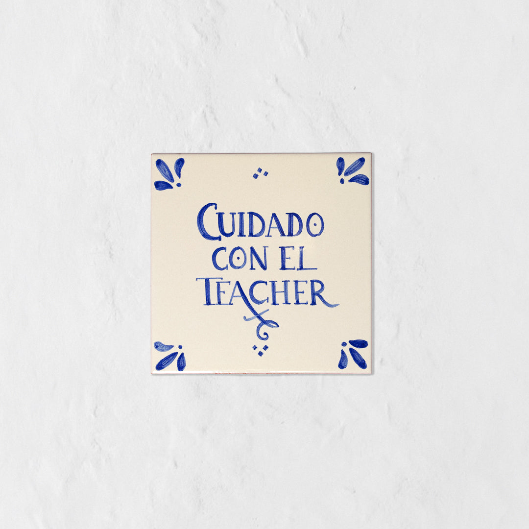 Azulejo decorativo de cerámica blanca con bordes y letras pintadas a mano en azul cobalto. En el centro se lee la frase en español 'CUIDADO CON EL TEACHER'. El diseño incluye detalles ornamentales en las esquinas y pequeños puntos decorativos, sobre un fondo de pared blanca con textura rugosa.