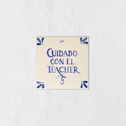 Azulejo decorativo de cerámica blanca con bordes y letras pintadas a mano en azul cobalto. En el centro se lee la frase en español 'CUIDADO CON EL TEACHER'. El diseño incluye detalles ornamentales en las esquinas y pequeños puntos decorativos, sobre un fondo de pared blanca con textura rugosa.