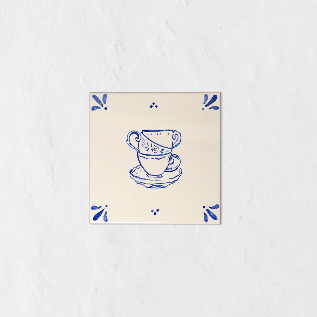 Azulejo decorativo de cerámica blanca con bordes y letras pintadas a mano en azul cobalto. En el centro tiene una ilustración de tres tazas de té. El diseño incluye detalles ornamentales en las esquinas y pequeños puntos decorativos, sobre un fondo de pared blanca con textura rugosa.