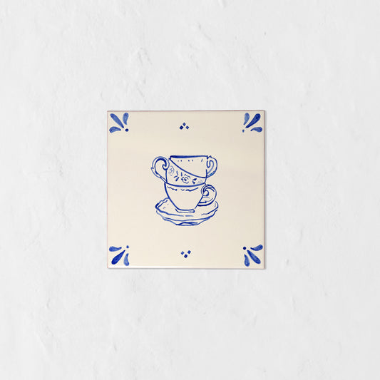 Azulejo decorativo de cerámica blanca con bordes y letras pintadas a mano en azul cobalto. En el centro tiene una ilustración de tres tazas de té. El diseño incluye detalles ornamentales en las esquinas y pequeños puntos decorativos, sobre un fondo de pared blanca con textura rugosa.