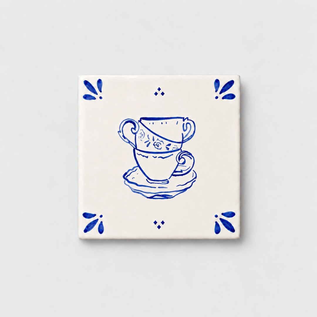 Azulejo decorativo de cerámica blanca con bordes y letras pintadas a mano en azul cobalto. En el centro tiene una ilustración de tres tazas de té. El diseño incluye detalles ornamentales en las esquinas y pequeños puntos decorativos, sobre un fondo de pared blanca con textura rugosa.