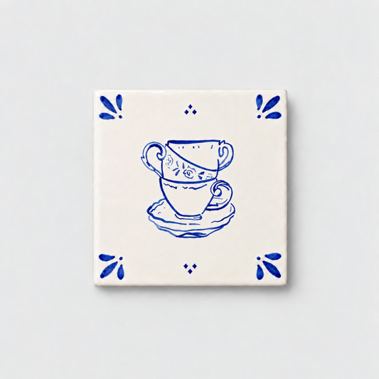 Azulejo decorativo de cerámica blanca con bordes y letras pintadas a mano en azul cobalto. En el centro tiene una ilustración de tres tazas de té. El diseño incluye detalles ornamentales en las esquinas y pequeños puntos decorativos, sobre un fondo de pared blanca con textura rugosa.