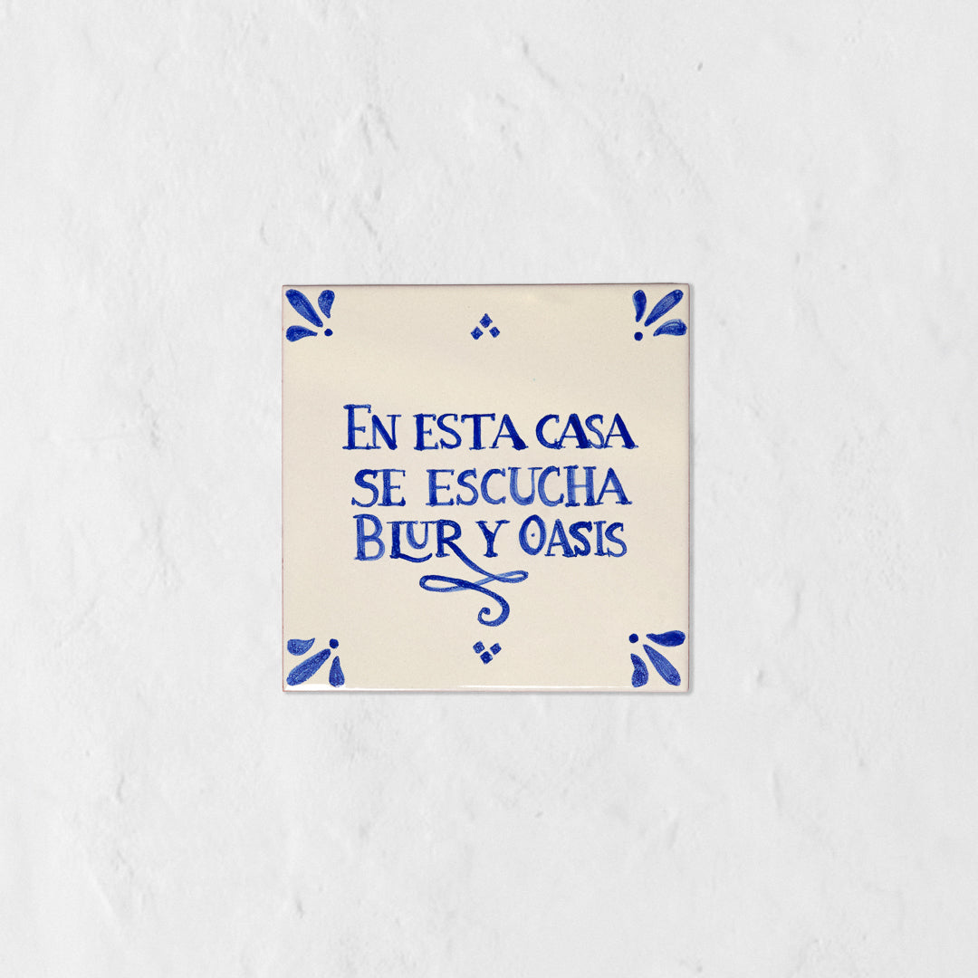 Azulejo decorativo de cerámica blanca con bordes y letras pintadas a mano en azul cobalto. En el centro se lee la frase en español 'EN ESTA CASA SE ESCUCHA BLUR Y OASIS'. El diseño incluye detalles ornamentales en las esquinas y pequeños puntos decorativos, sobre un fondo de pared blanca con textura rugosa.