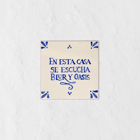 Azulejo decorativo de cerámica blanca con bordes y letras pintadas a mano en azul cobalto. En el centro se lee la frase en español 'EN ESTA CASA SE ESCUCHA BLUR Y OASIS'. El diseño incluye detalles ornamentales en las esquinas y pequeños puntos decorativos, sobre un fondo de pared blanca con textura rugosa.