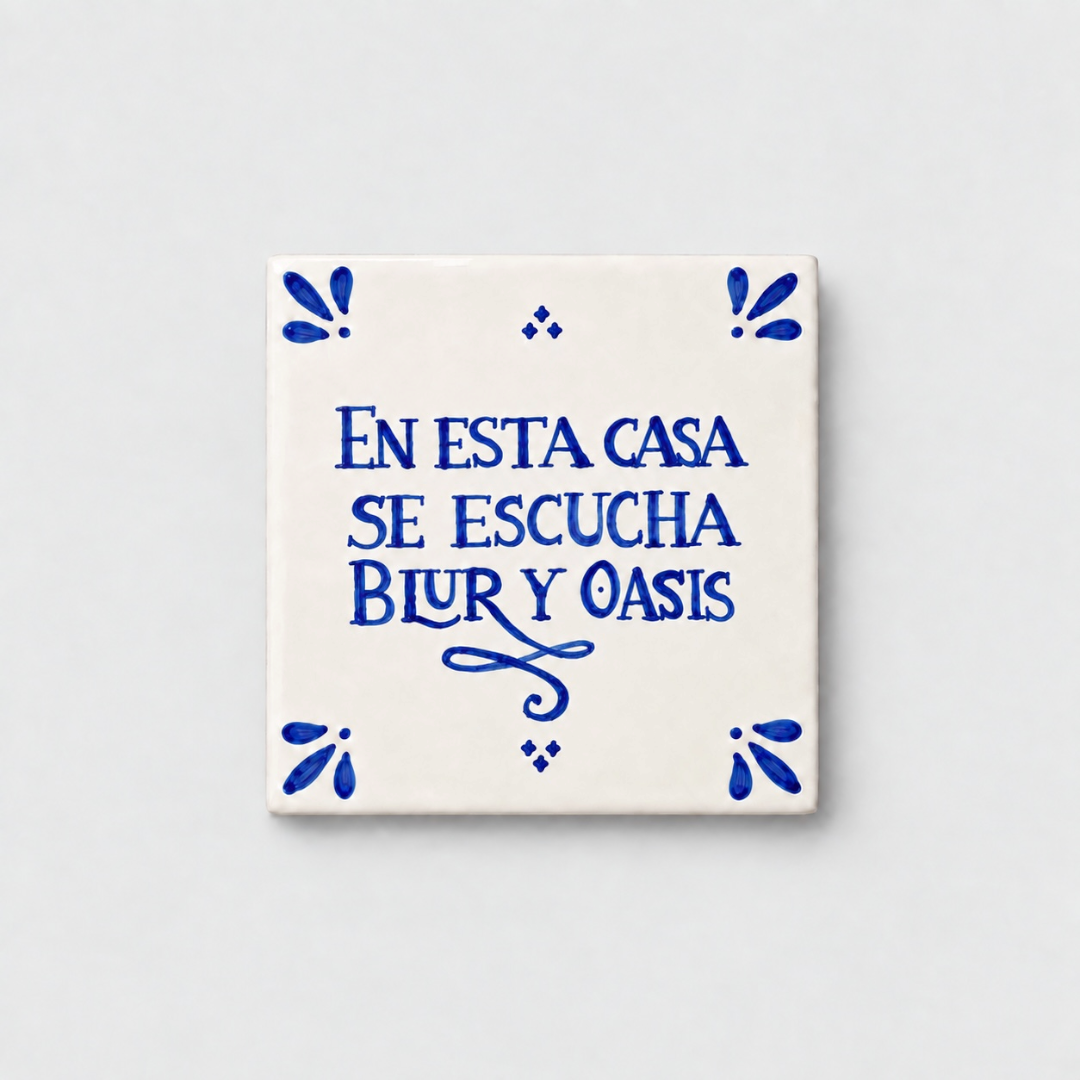 Azulejo decorativo de cerámica blanca con bordes y letras pintadas a mano en azul cobalto. En el centro se lee la frase en español 'EN ESTA CASA SE ESCUCHA BLUR Y OASIS'. El diseño incluye detalles ornamentales en las esquinas y pequeños puntos decorativos, sobre un fondo de pared blanca con textura rugosa.