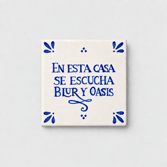 Azulejo decorativo de cerámica blanca con bordes y letras pintadas a mano en azul cobalto. En el centro se lee la frase en español 'EN ESTA CASA SE ESCUCHA BLUR Y OASIS'. El diseño incluye detalles ornamentales en las esquinas y pequeños puntos decorativos, sobre un fondo de pared blanca con textura rugosa.