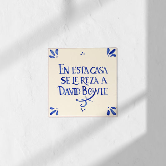 Azulejo decorativo de cerámica blanca con bordes y letras pintadas a mano en azul cobalto. En el centro se lee la frase en español 'EN ESTA CASA SE LE REZA A DAVID BOWIE'. El diseño incluye detalles ornamentales en las esquinas y pequeños puntos decorativos, sobre un fondo de pared blanca con textura rugosa.