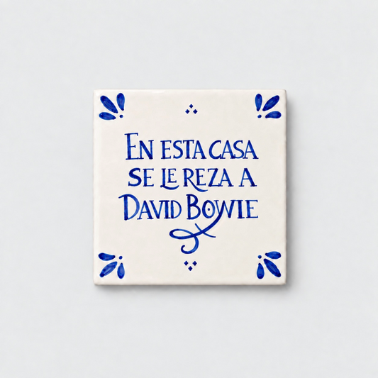 Azulejo decorativo de cerámica blanca con bordes y letras pintadas a mano en azul cobalto. En el centro se lee la frase en español 'EN ESTA CASA SE LE REZA A DAVID BOWIE'. El diseño incluye detalles ornamentales en las esquinas y pequeños puntos decorativos, sobre un fondo de pared blanca con textura rugosa.