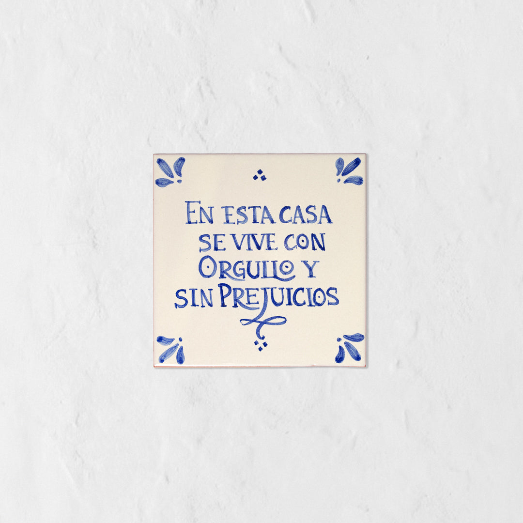 Azulejo decorativo de cerámica blanca con bordes y letras pintadas a mano en azul cobalto. En el centro se lee la frase en español 'EN ESTA CASA SE VIVE CON ORGULLO Y SIN PREJUICIOS'. El diseño incluye detalles ornamentales en las esquinas y pequeños puntos decorativos, sobre un fondo de pared blanca con textura rugosa.