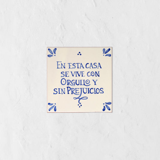 Azulejo decorativo de cerámica blanca con bordes y letras pintadas a mano en azul cobalto. En el centro se lee la frase en español 'EN ESTA CASA SE VIVE CON ORGULLO Y SIN PREJUICIOS'. El diseño incluye detalles ornamentales en las esquinas y pequeños puntos decorativos, sobre un fondo de pared blanca con textura rugosa.