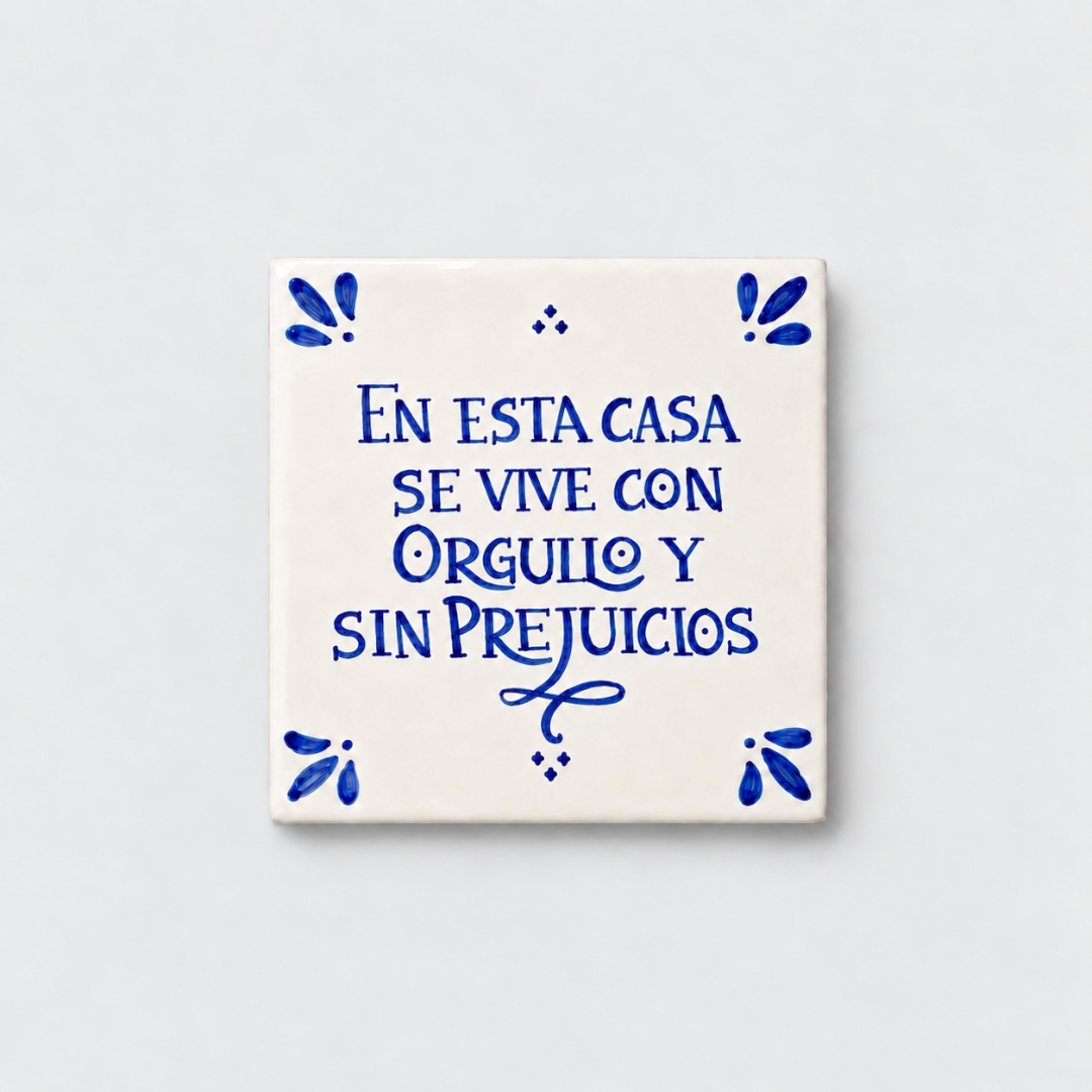 Azulejo decorativo de cerámica blanca con bordes y letras pintadas a mano en azul cobalto. En el centro se lee la frase en español 'EN ESTA CASA SE VIVE CON ORGULLO Y SIN PREJUICIOS'. El diseño incluye detalles ornamentales en las esquinas y pequeños puntos decorativos, sobre un fondo de pared blanca con textura rugosa.