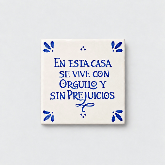 Azulejo decorativo de cerámica blanca con bordes y letras pintadas a mano en azul cobalto. En el centro se lee la frase en español 'EN ESTA CASA SE VIVE CON ORGULLO Y SIN PREJUICIOS'. El diseño incluye detalles ornamentales en las esquinas y pequeños puntos decorativos, sobre un fondo de pared blanca con textura rugosa.