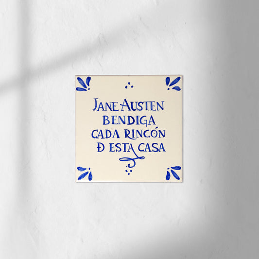 Azulejo decorativo de cerámica blanca con bordes y letras pintadas a mano en azul cobalto. En el centro se lee la frase en español 'JANE AUSTEN BENDIGA CADA RINCÓN DE ESTA CASA'. El diseño incluye detalles ornamentales en las esquinas y pequeños puntos decorativos, sobre un fondo de pared blanca con textura rugosa.