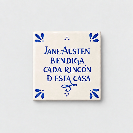 Azulejo decorativo de cerámica blanca con bordes y letras pintadas a mano en azul cobalto. En el centro se lee la frase en español 'JANE AUSTEN BENDIGA CADA RINCÓN DE ESTA CASA'. El diseño incluye detalles ornamentales en las esquinas y pequeños puntos decorativos, sobre un fondo de pared blanca con textura rugosa.