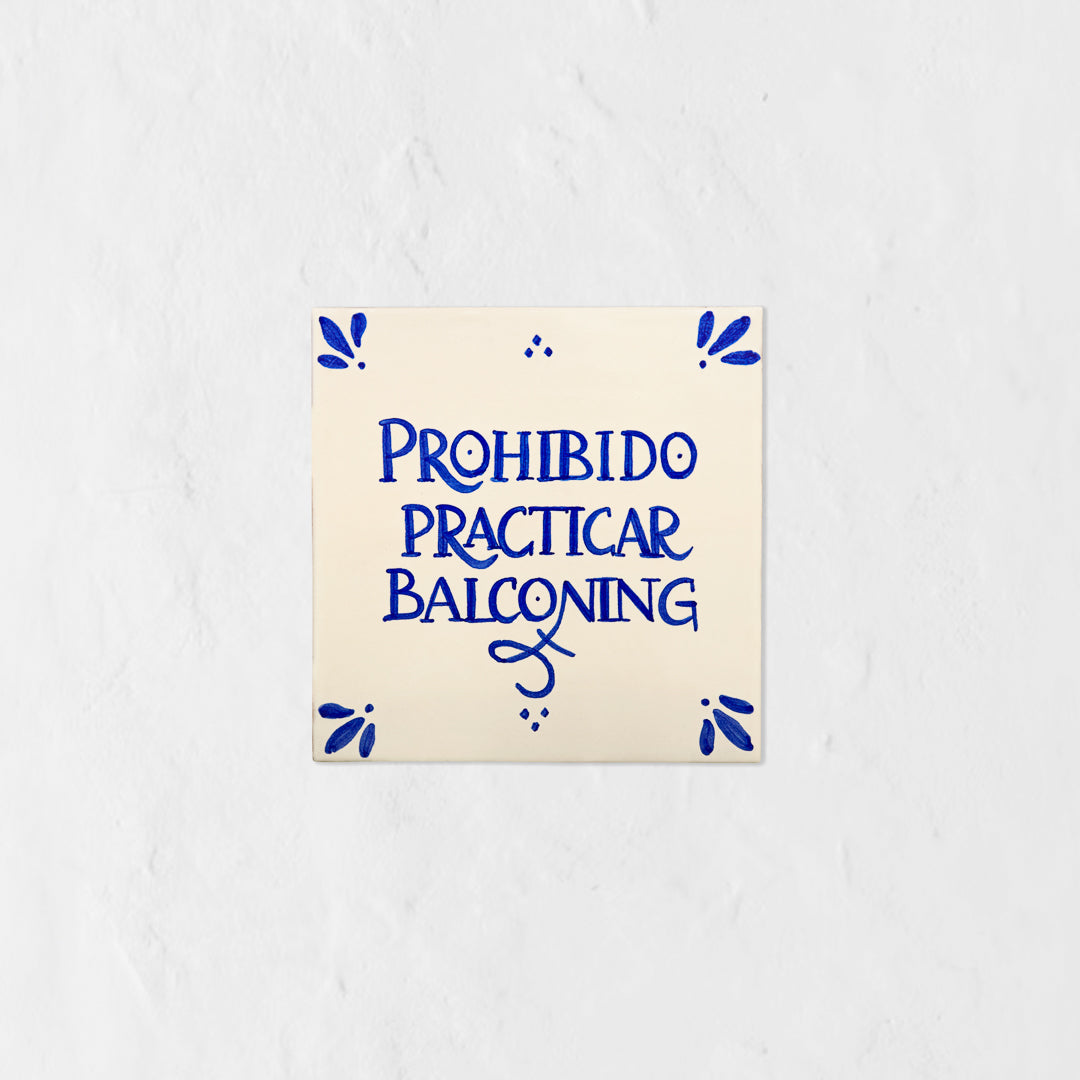 Azulejo decorativo de cerámica blanca con bordes y letras pintadas a mano en azul cobalto. En el centro se lee la frase en español 'PROHIBIDO PRACTICAR BALCONING'. El diseño incluye detalles ornamentales en las esquinas y pequeños puntos decorativos, sobre un fondo de pared blanca con textura rugosa.