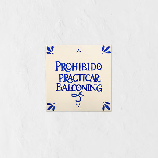 Azulejo decorativo de cerámica blanca con bordes y letras pintadas a mano en azul cobalto. En el centro se lee la frase en español 'PROHIBIDO PRACTICAR BALCONING'. El diseño incluye detalles ornamentales en las esquinas y pequeños puntos decorativos, sobre un fondo de pared blanca con textura rugosa.