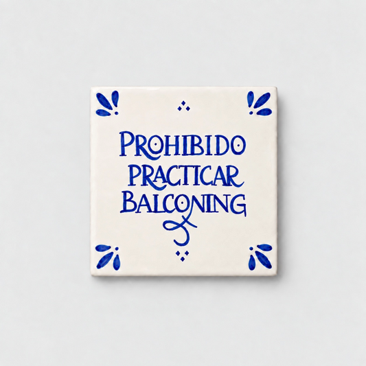 Azulejo decorativo de cerámica blanca con bordes y letras pintadas a mano en azul cobalto. En el centro se lee la frase en español 'PROHIBIDO PRACTICAR BALCONING'. El diseño incluye detalles ornamentales en las esquinas y pequeños puntos decorativos, sobre un fondo de pared blanca con textura rugosa.