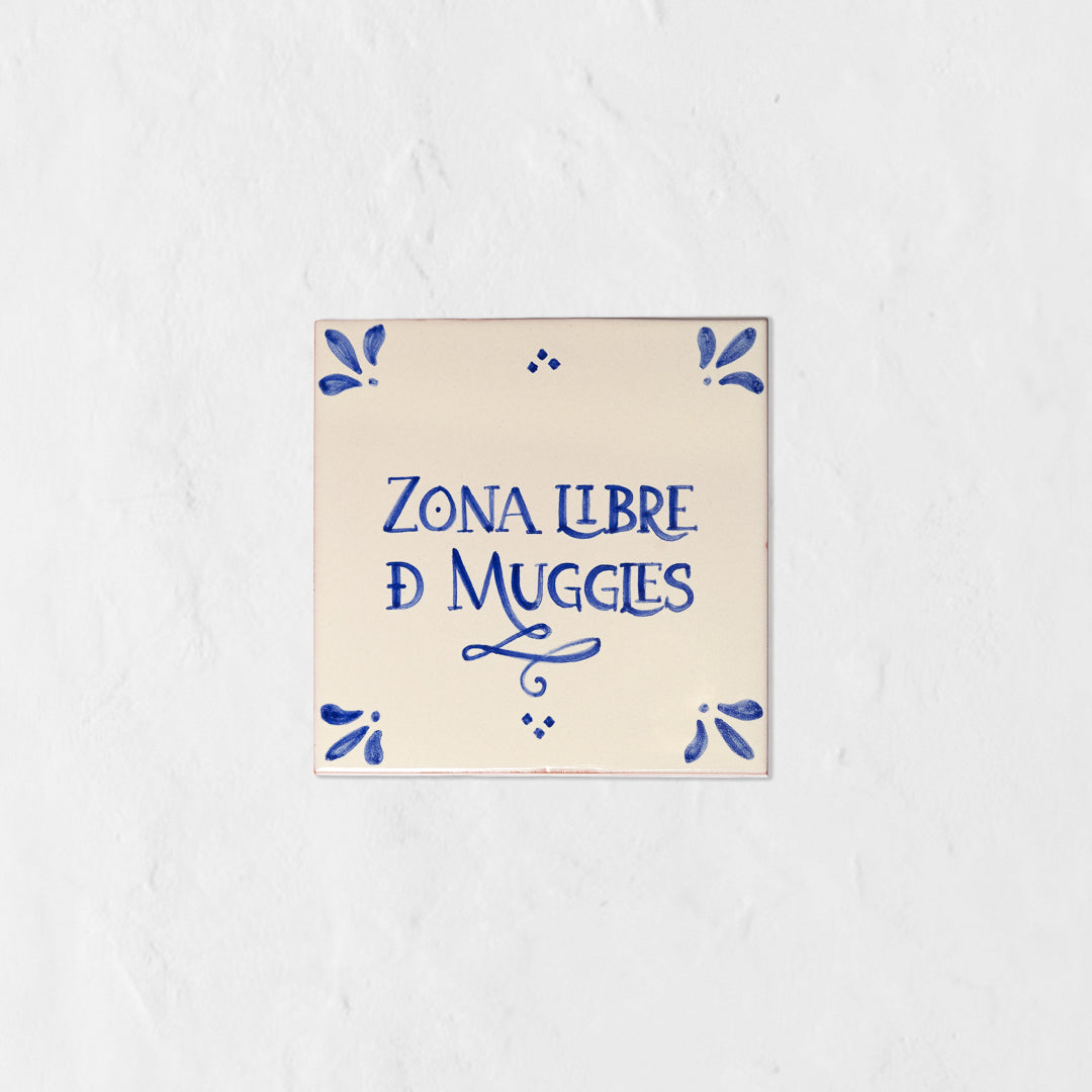 Azulejo decorativo de cerámica blanca con bordes y letras pintadas a mano en azul cobalto. En el centro se lee la frase en español 'ZONA LIBRE DE MUGGLES'. El diseño incluye detalles ornamentales en las esquinas y pequeños puntos decorativos, sobre un fondo de pared blanca con textura rugosa.
