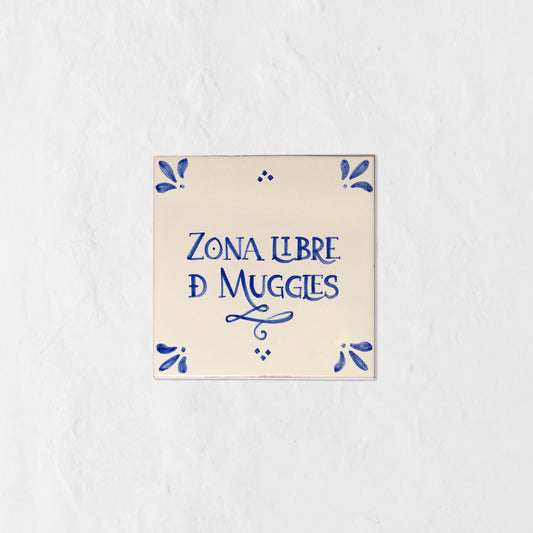 Azulejo decorativo de cerámica blanca con bordes y letras pintadas a mano en azul cobalto. En el centro se lee la frase en español 'ZONA LIBRE DE MUGGLES'. El diseño incluye detalles ornamentales en las esquinas y pequeños puntos decorativos, sobre un fondo de pared blanca con textura rugosa.