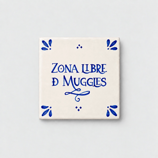 Azulejo decorativo de cerámica blanca con bordes y letras pintadas a mano en azul cobalto. En el centro se lee la frase en español 'ZONA LIBRE DE MUGGLES'. El diseño incluye detalles ornamentales en las esquinas y pequeños puntos decorativos, sobre un fondo de pared blanca con textura rugosa.
