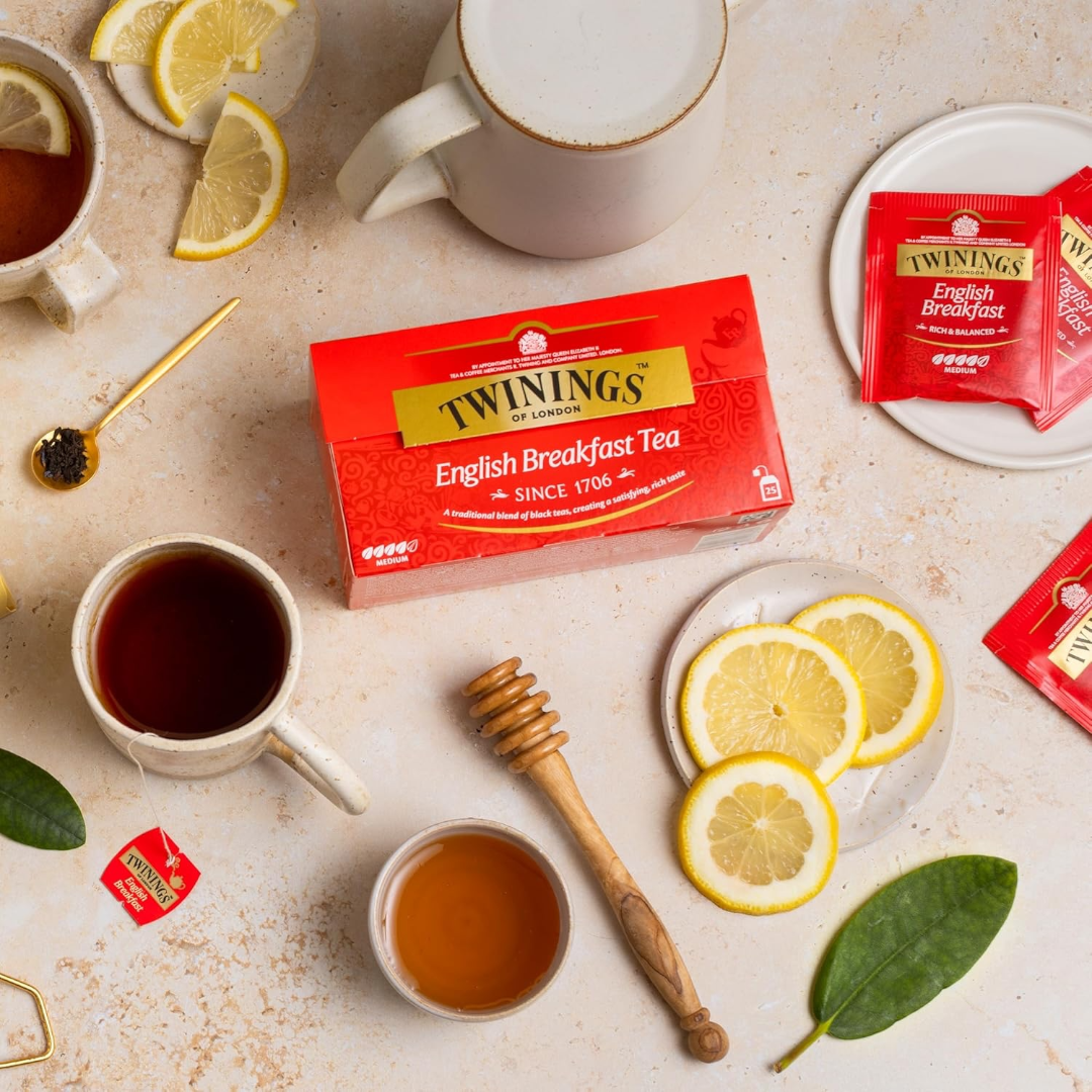 Caja de té Twinings English Breakfast rodeada de tazas de té caliente, rodajas de limón fresco, miel con un dispensador de madera y bolsitas individuales sobre un fondo texturizado claro.