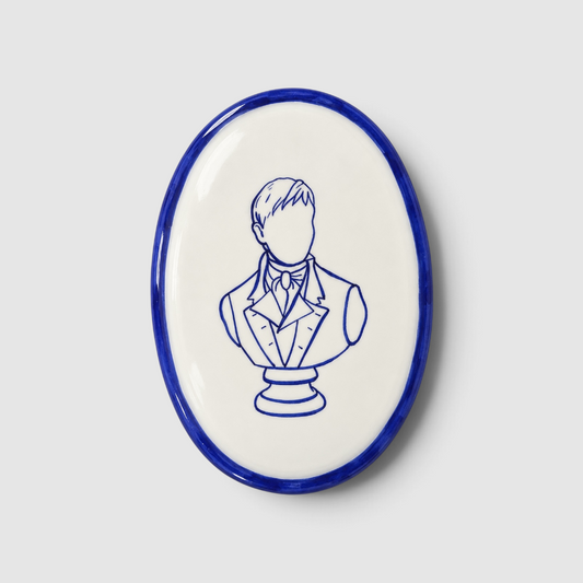 Placa ovalada de cerámica artesanal «Mr Darcy no existe» de Superbritánico, con ilustración del busto de Mr Darcy y borde pintados en azul cobalto sobre fondo blanco.