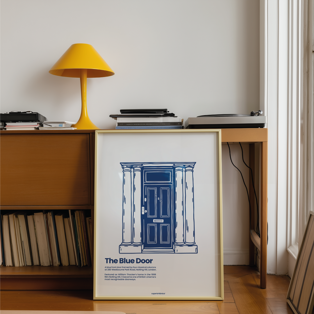 Póster The Blue Door de Superbritánico, inspirado en la icónica puerta azul de Notting Hill, presentado en un marco fino dorado apoyado sobre un mueble de madera. La escena está ambientada como un rincón de interiorismo de estilo retro y editorial, con una lámpara amarilla, libros y revistas apilados, un tocadiscos y discos de vinilo en la balda inferior, junto a una gran ventana que aporta luz natural.