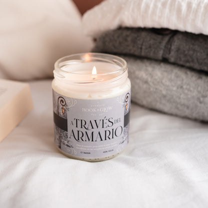 Vela de Book and Glow A través del armario encendida, presentada en recipiente de cristal, etiqueta gris con título en color negro sobre colcha con mantas.