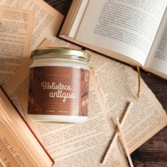 Vela de Book and Glow Biblioteca antigua presentada en recipiente de cristal cerrado, etiqueta marrón con título en color blanco sobre mesa con libros.