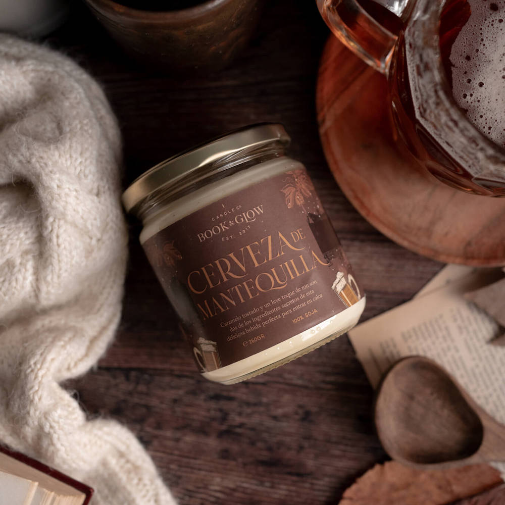 Vela de book and Glow Cerveza de mantequilla presentada en recipiente de cristal, etiqueta marrón con título en naranja sobre mesa de madera decorada con jarras de cerveza.