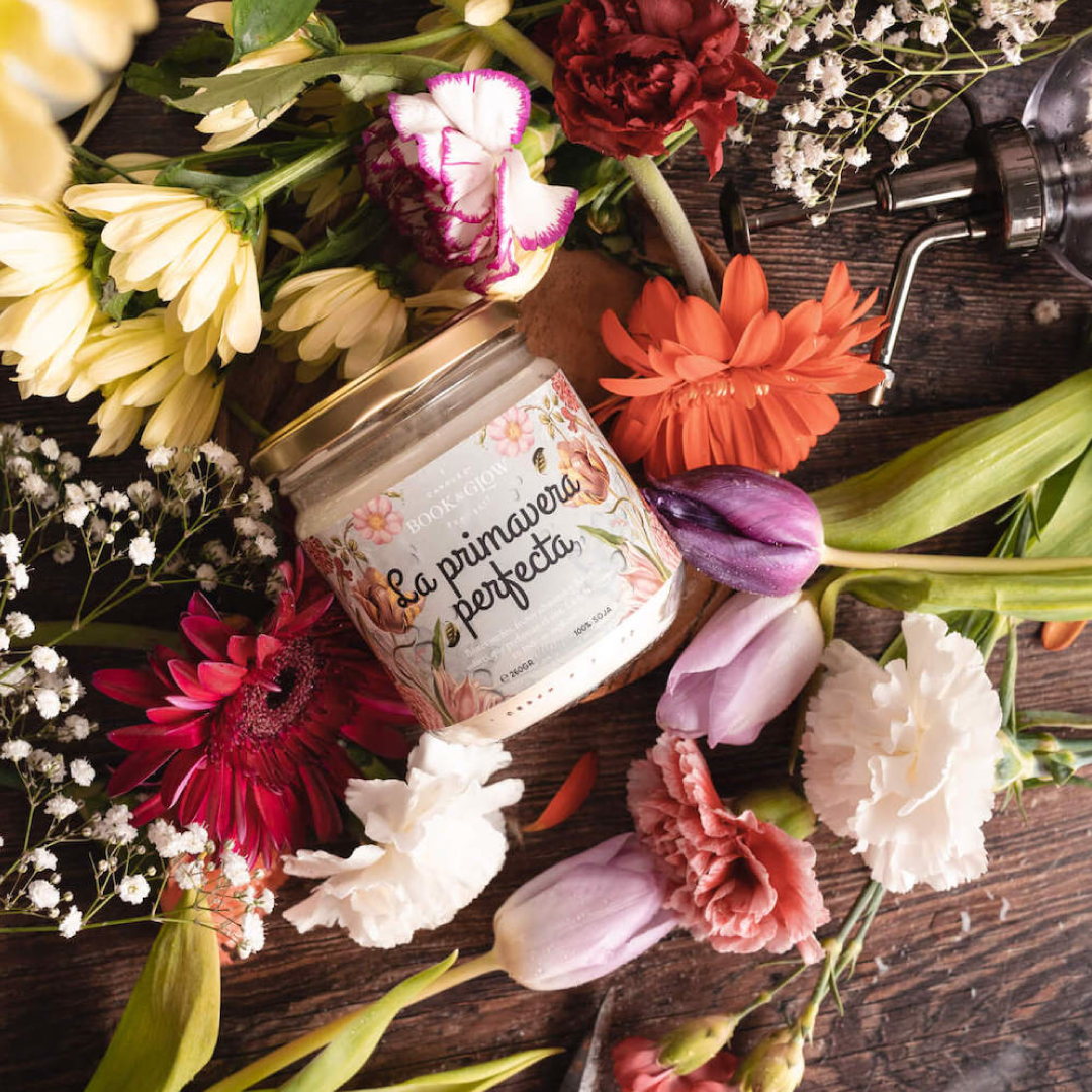 Vela La primavera perfecta de Book and Glow presentada en tarro de cristal cerrado junto a bodegón de flores. La etiqueta de la vela es celeste con ilustraciones florales.