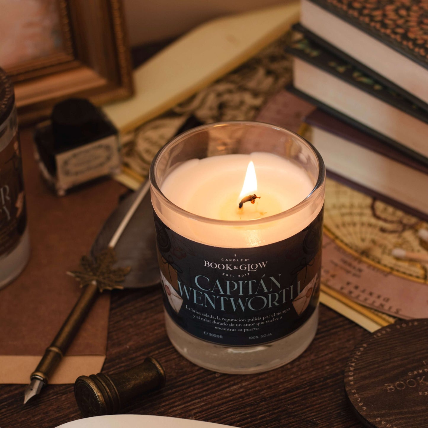 vela del Capitán Wentworth de Book and glow encendida, junto a libros y pluma, sobre escritorio de madera.