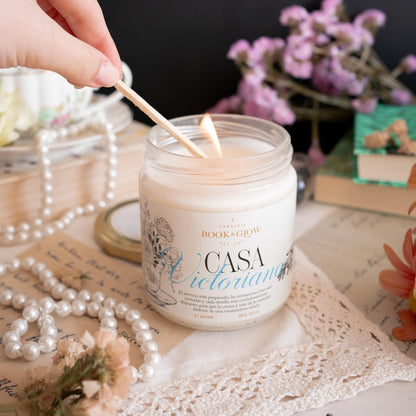 Vela de Book and Glow Casa Victoriana siendo encendida, presentada en recipiente de cristal con etiqueta blanca y título en negro y celeste, sobre mesa victoriana con collar de perlas y flores color lavanda.