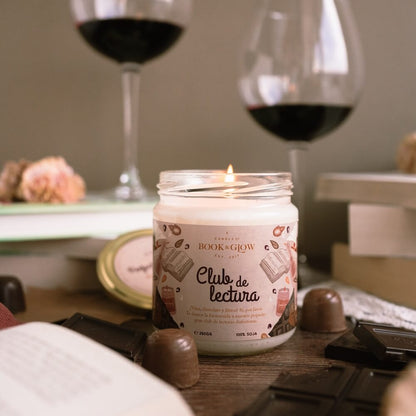 Vela de Book and Glow Club de lectura presentada en recipiente de cristal, etiqueta blanca con título en color negro sobre mesa con bombones y copas de vino.