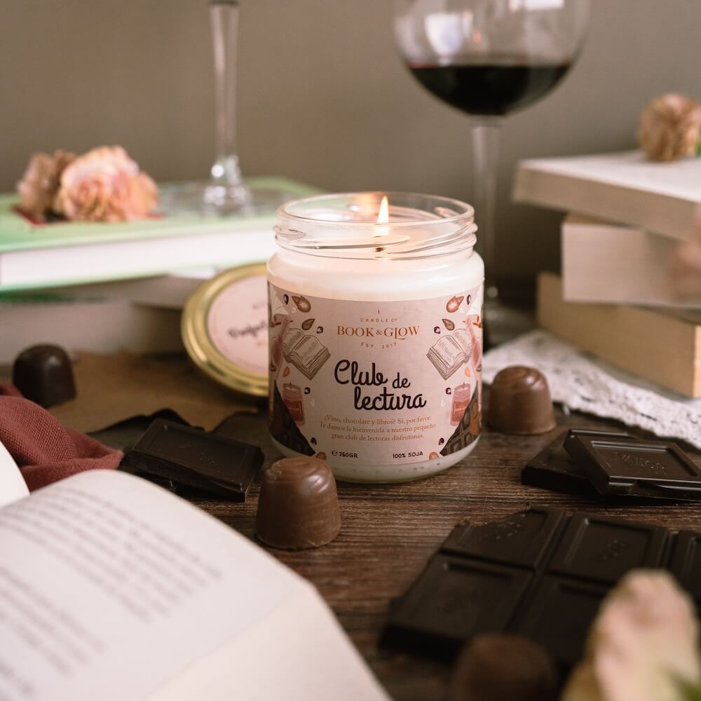 Vela de Book and Glow Club de lectura encendida presentada en recipiente de cristal, etiqueta blanca con título en color negro sobre mesa con bombones y copas de vino.