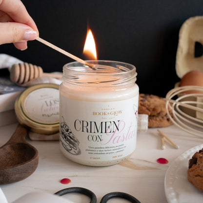 Vela de Book and Glow Crimen con pastas presentada en recipiente de cristal con etiqueta blanca y título en negro y rosa, siendo encendida con una cerilla.
