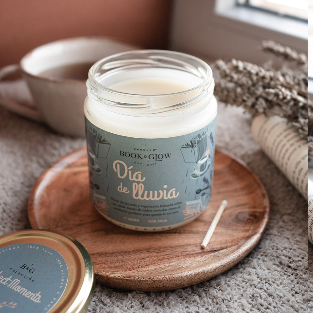 Vela de book and Glow Día de lluvia presentada en recipiente de cristal, etiqueta celeste con título en blanco sobre plato de madera y manta.