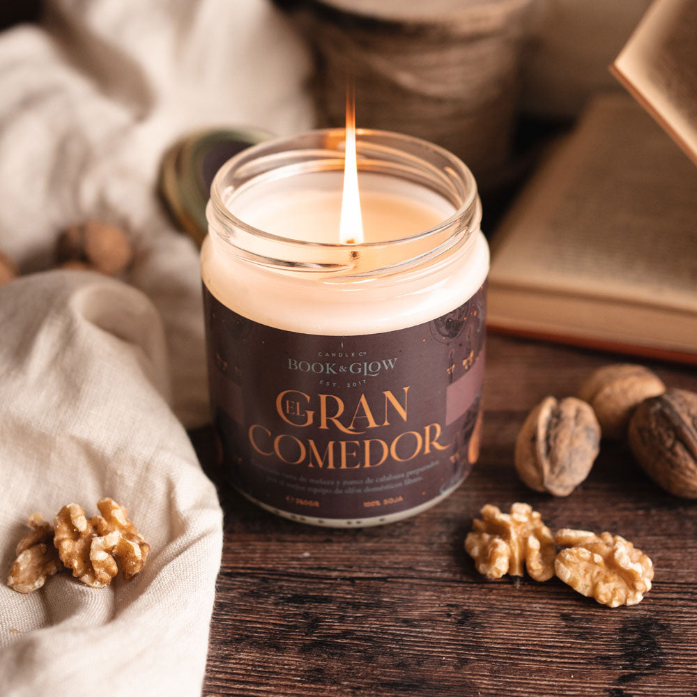 Vela de Book and Glow El Gran Comedor presentada en recipiente de cristal, etiqueta marrón con título en color calabaza sobre mesa con nueces.