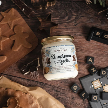 Vela de book and Glow El invierno perfecto presentada en recipiente de cristal, etiqueta blanca y celeste con título en negro sobre mesa de madera con decoración navideña.