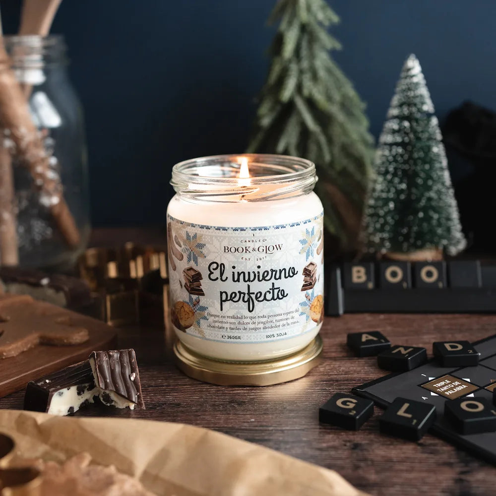 Vela de book and Glow El invierno perfecto encendida presentada en recipiente de cristal, etiqueta blanca y celeste con título en negro sobre mesa de madera con decoración navideña.