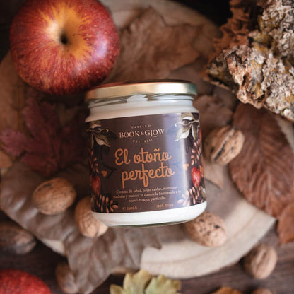 Vela de book and Glow El otoño perfecto presentada en recipiente de cristal cerrado, etiqueta marrón con título en naranja sobre hojas secas y junto a una manzana.