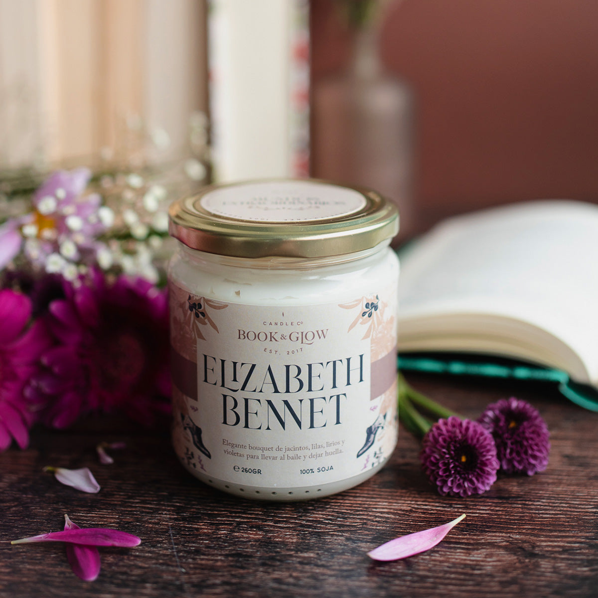 Vela de Book and Glow Elizabeth bennet, presentada en recipiente de cristal, etiqueta clara con título en color negro sobre mesa con flores y libros.